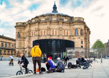 Young adults play with bikes and skateboards on Bristo Square with McEwan Hall of the University of Edinburgh in the background, in downtown Edinburgh, Scotland, UK on an overcast day.